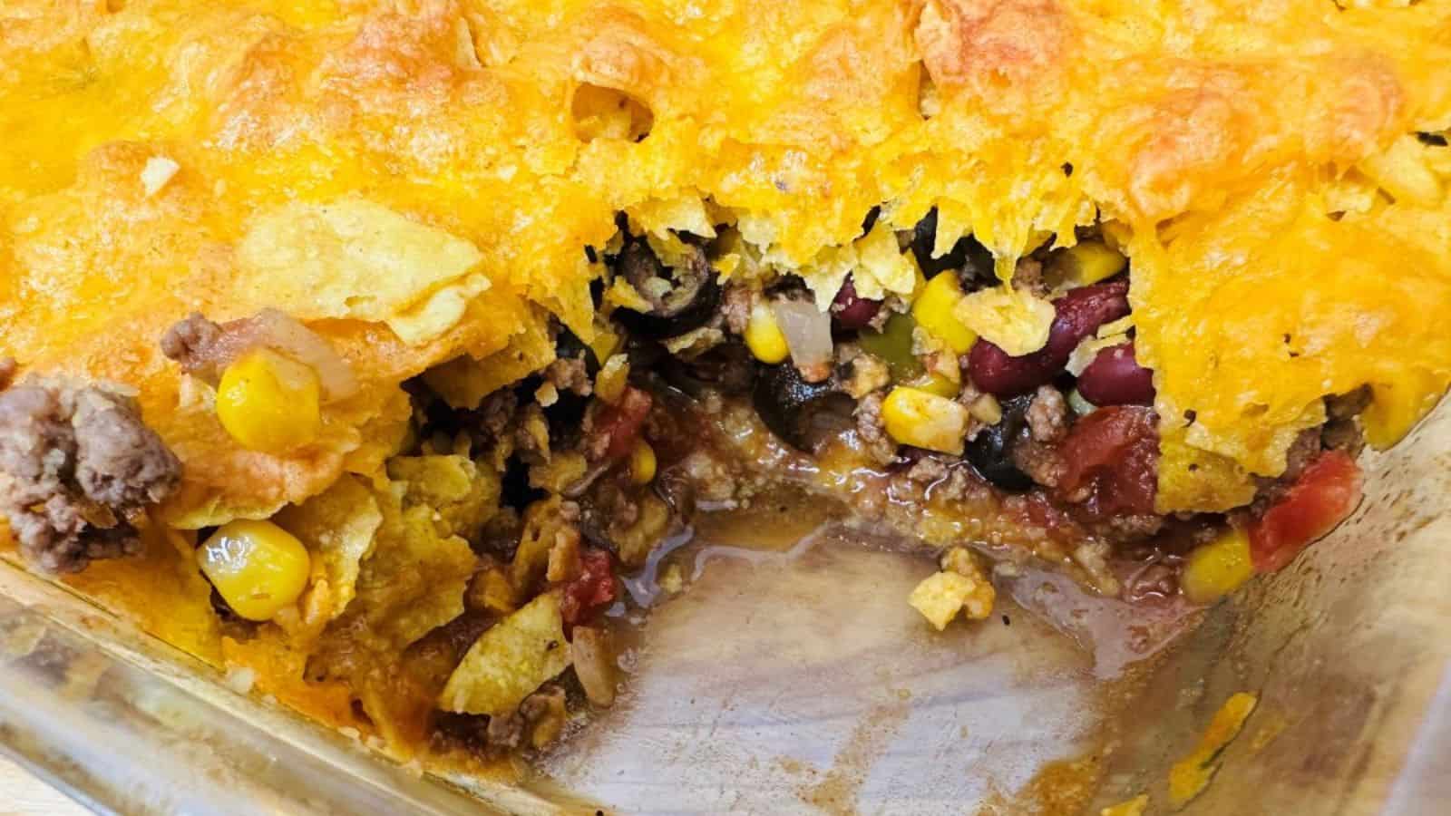 A partially served Mexican casserole in a baking dish, showing layers of corn, beans, cheese, and ground meat.