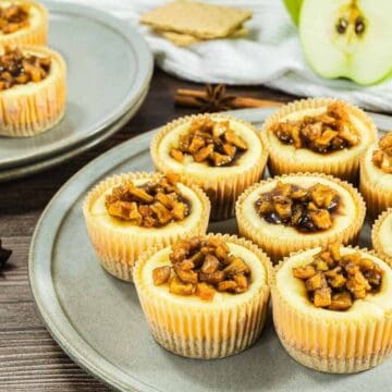 Mini cheesecakes topped with caramelized apples on plates, with a green apple and graham crackers in the background.