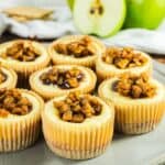 Mini cheesecakes topped with caramelized apple pieces arranged on a plate, with apples in the background.