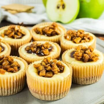 Mini cheesecakes topped with caramelized apple pieces arranged on a plate, with apples in the background.