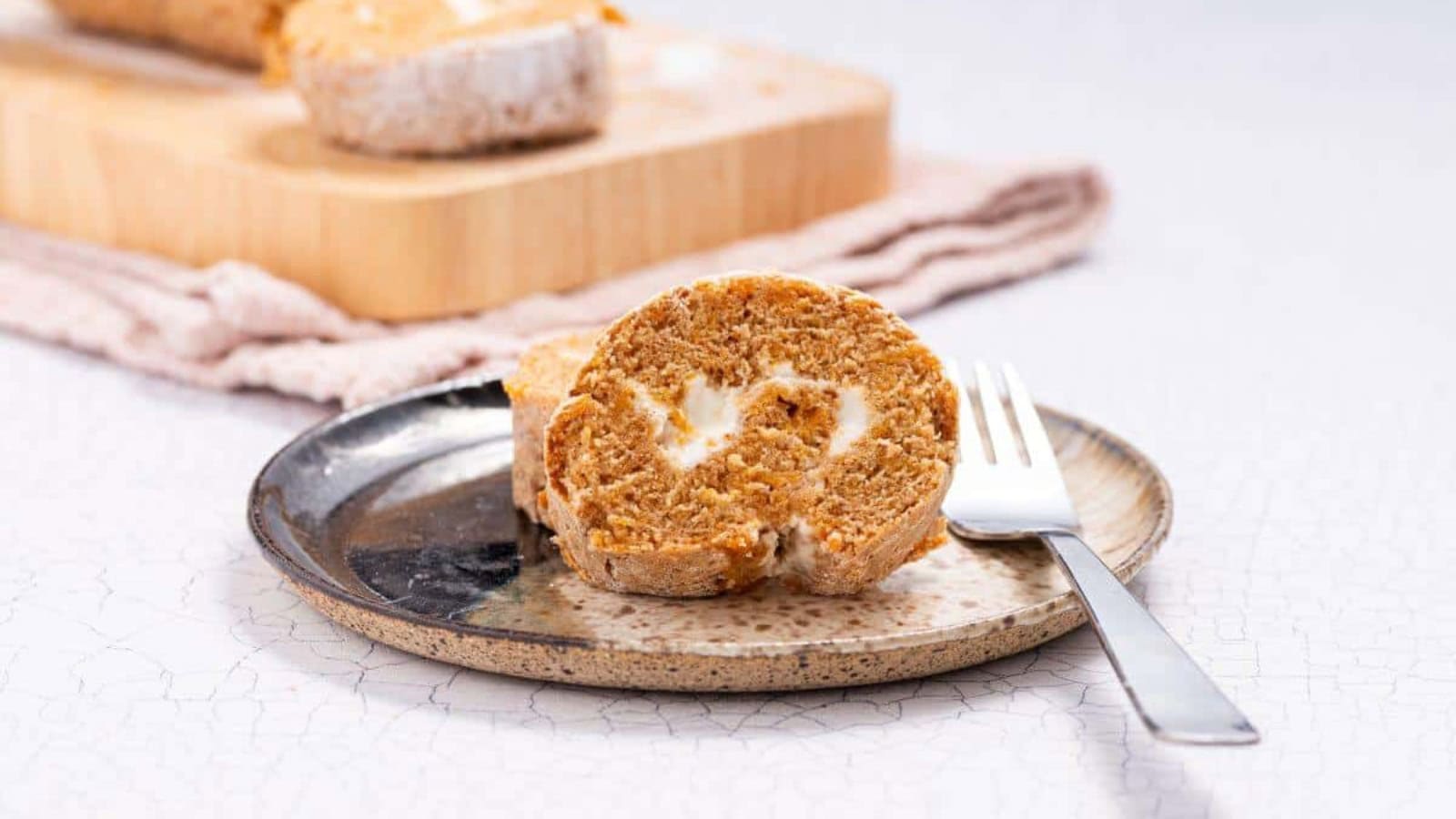 A slice of pumpkin roll cake with cream filling on a plate, fork beside it. More slices and a rolled cake are on a cutting board in the background.