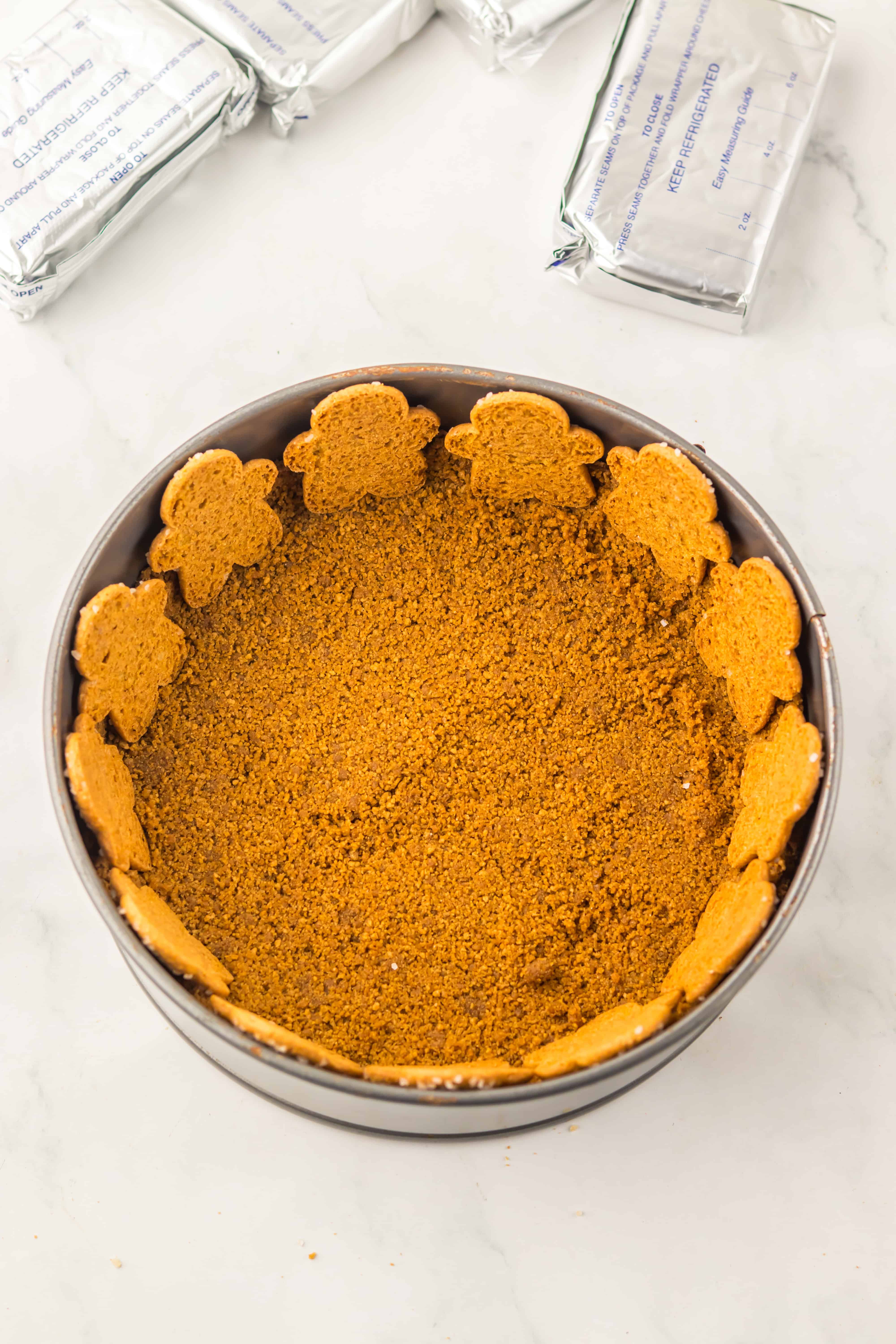 Round pan with a crumb crust and gingerbread cookies around the edge, perfect for creating a festive No Bake Gingerbread Cheesecake, with cream cheese packages nearby.