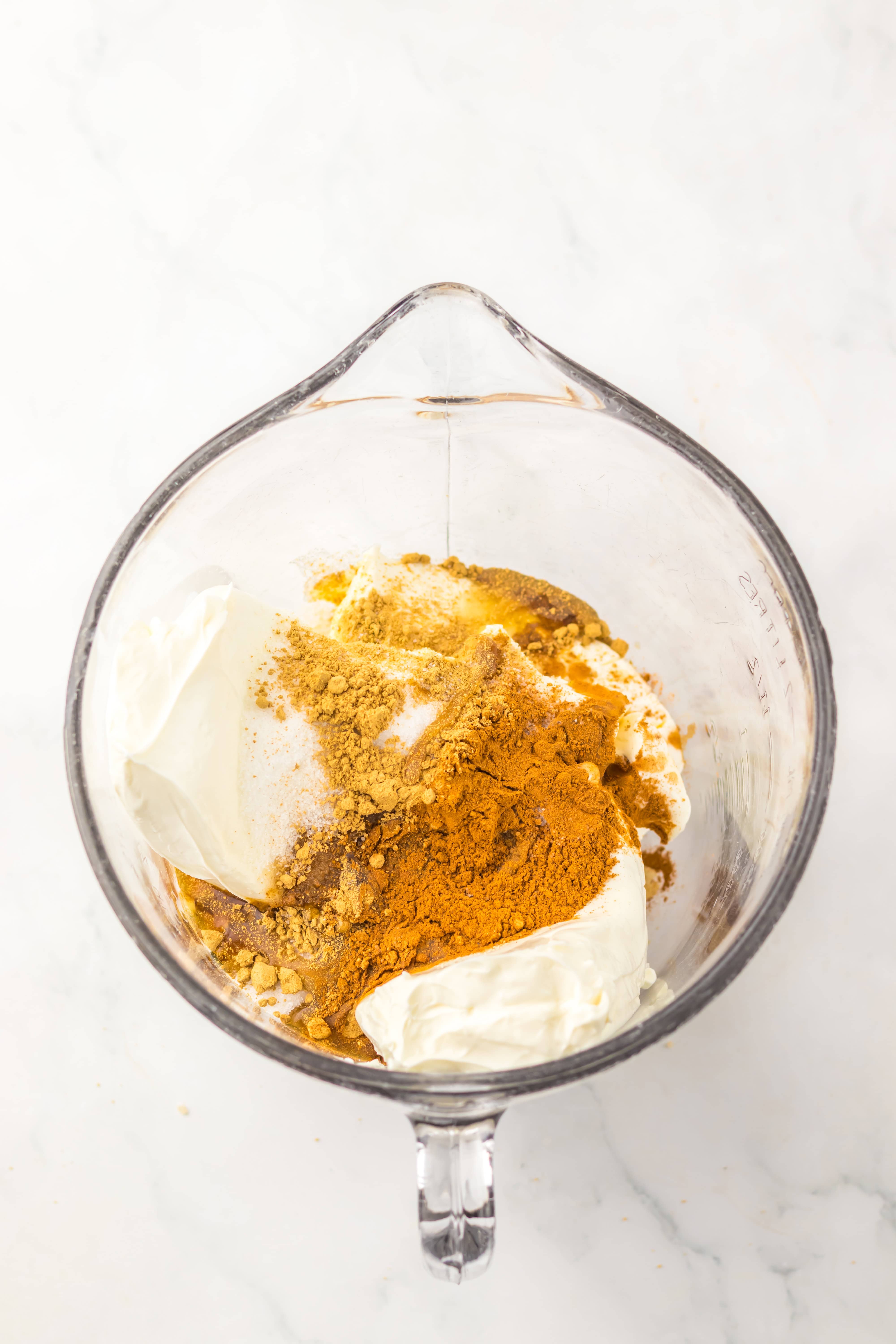 A glass bowl with food ingredients in it, ready to be mixed for a delicious No Bake Gingerbread Cheesecake.