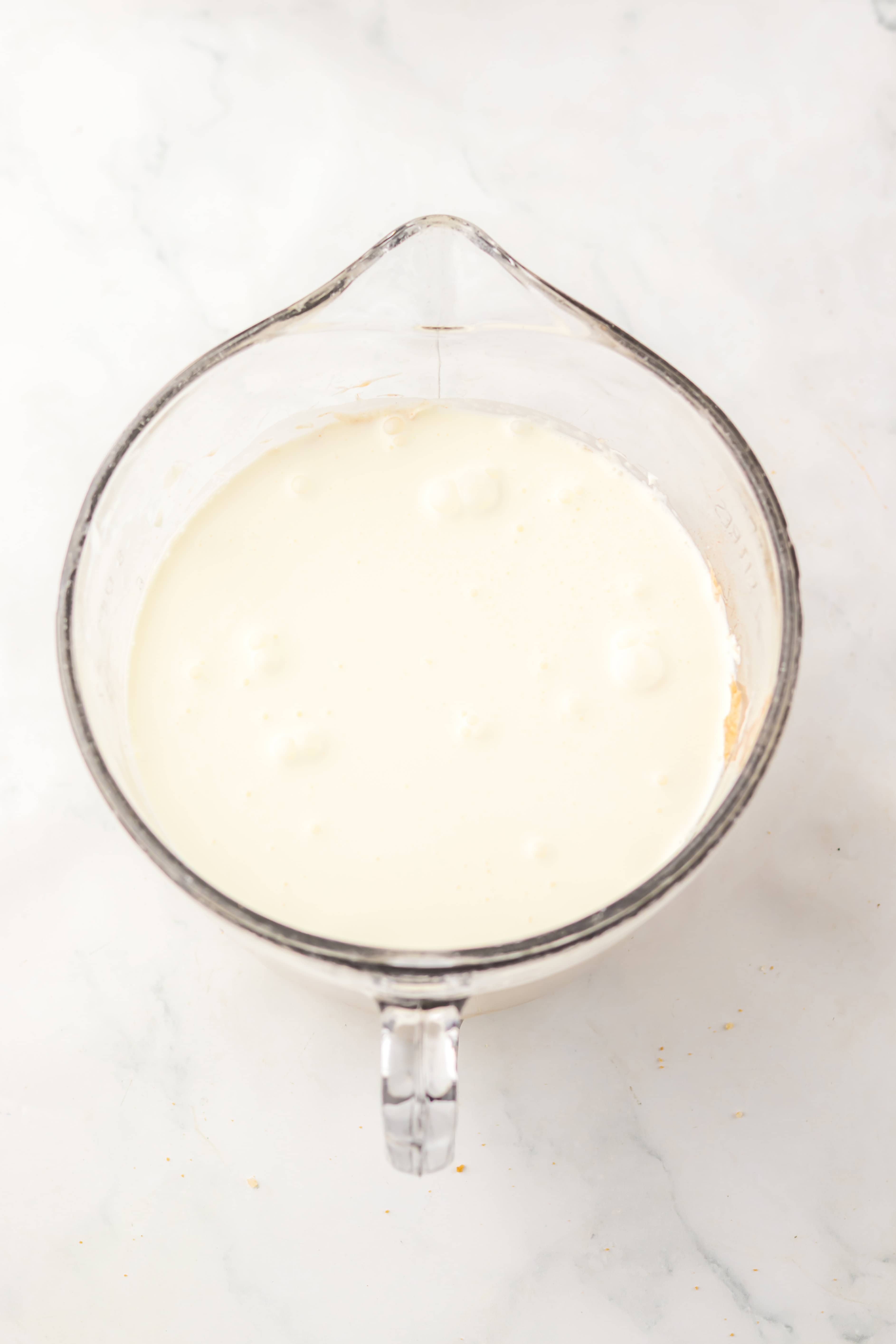 Glass measuring cup filled with thick white cream on a white marble surface, perfect for preparing a No Bake Gingerbread Cheesecake.