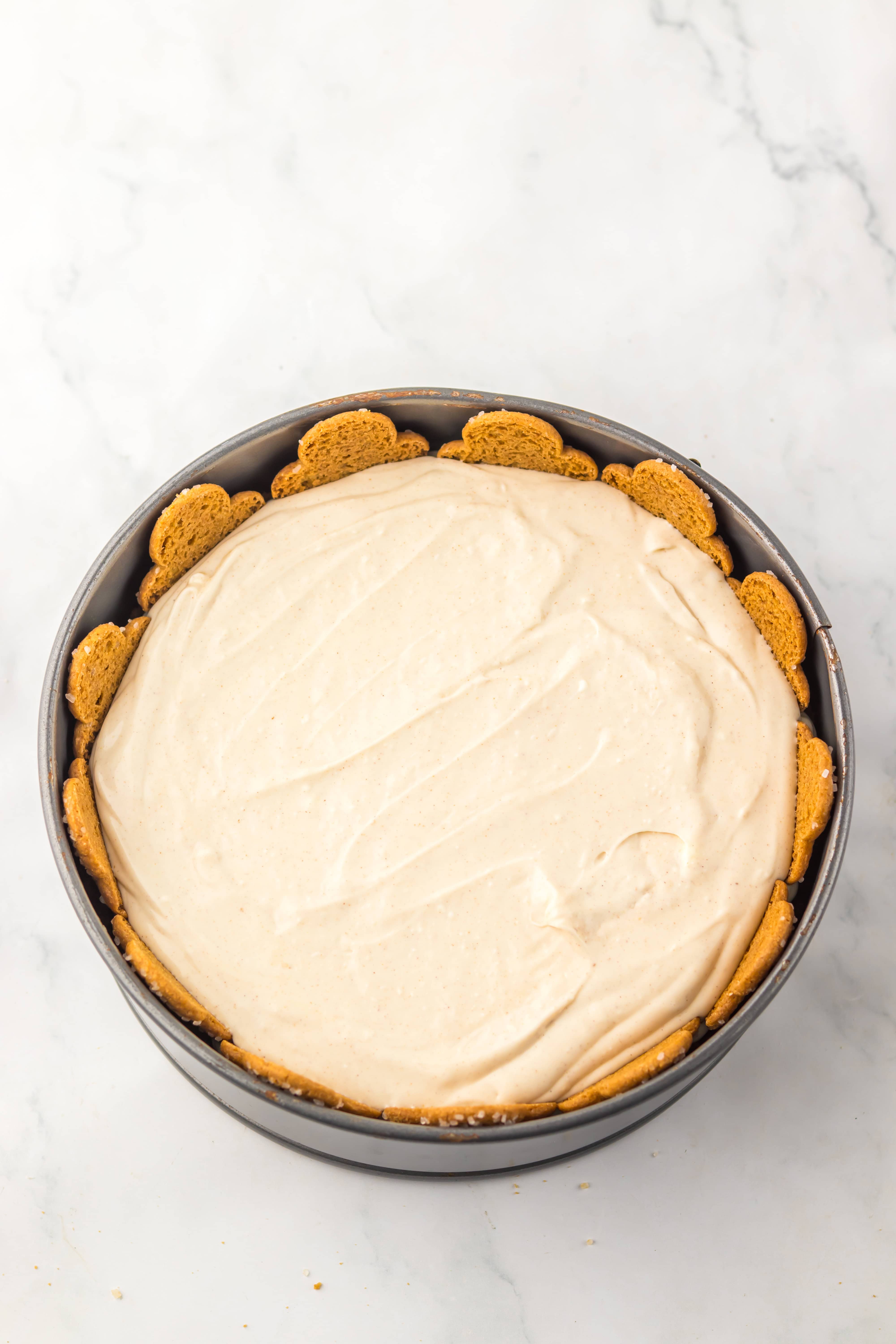 A No Bake Gingerbread Cheesecake in a round pan, lined with gingersnap cookies along the edge, sits on a marble surface.