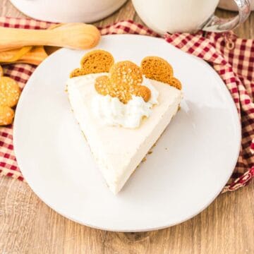 A slice of No Bake Gingerbread Cheesecake with whipped cream and cookies on top, served on a white plate with a red checkered cloth.