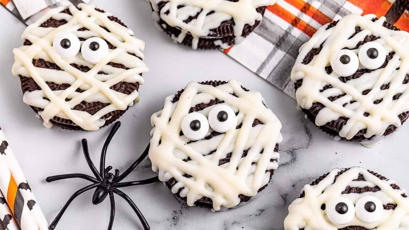 Mummy-themed Oreo cookies with white icing lines and candy eyes on a tabletop.
