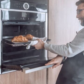 Man taking fresh baked bread out of an oven in a modern kitchen, wearing glasses and an apron.
