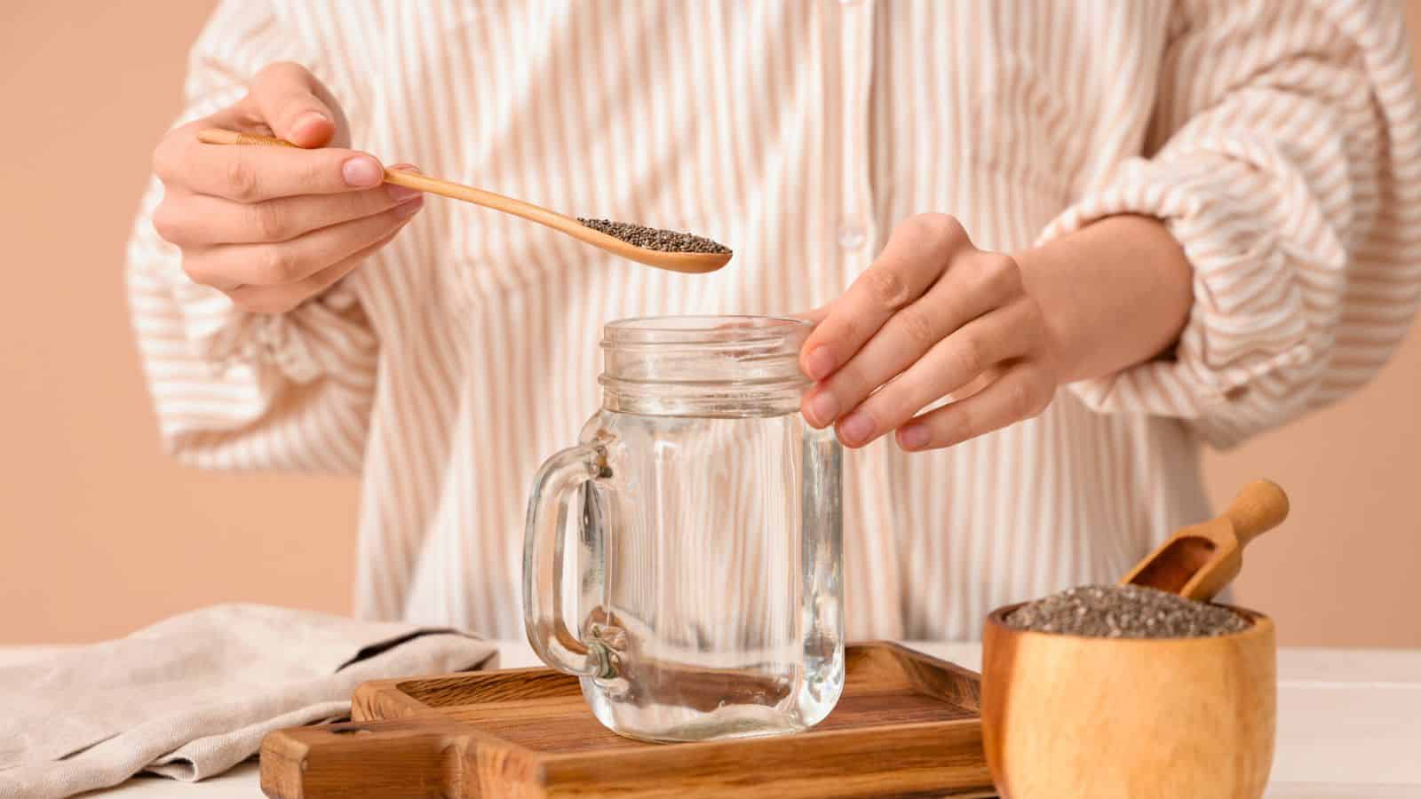 Person adding chia seeds from a spoon into a glass jar of water, with more seeds in a wooden bowl nearby.