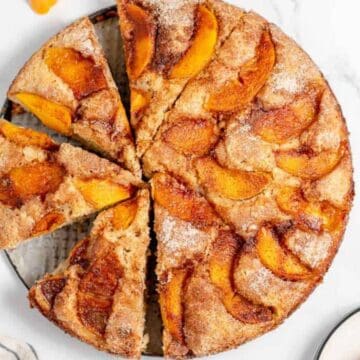 Round peach cake sliced into wedges, topped with cinnamon and peach slices, on a white background.