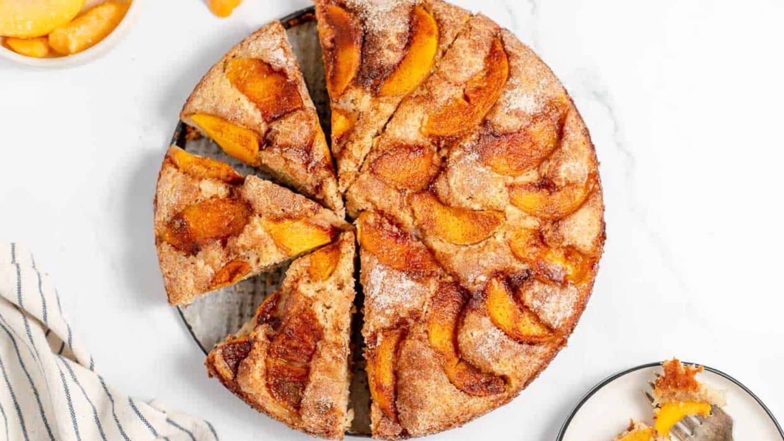 A sliced peach cobbler on a plate, with a slice served on a smaller plate. A striped napkin and a bowl of peach slices are placed nearby.