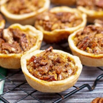 Mini pecan pies cooling on a wire rack, with a green cloth and pecans scattered nearby.