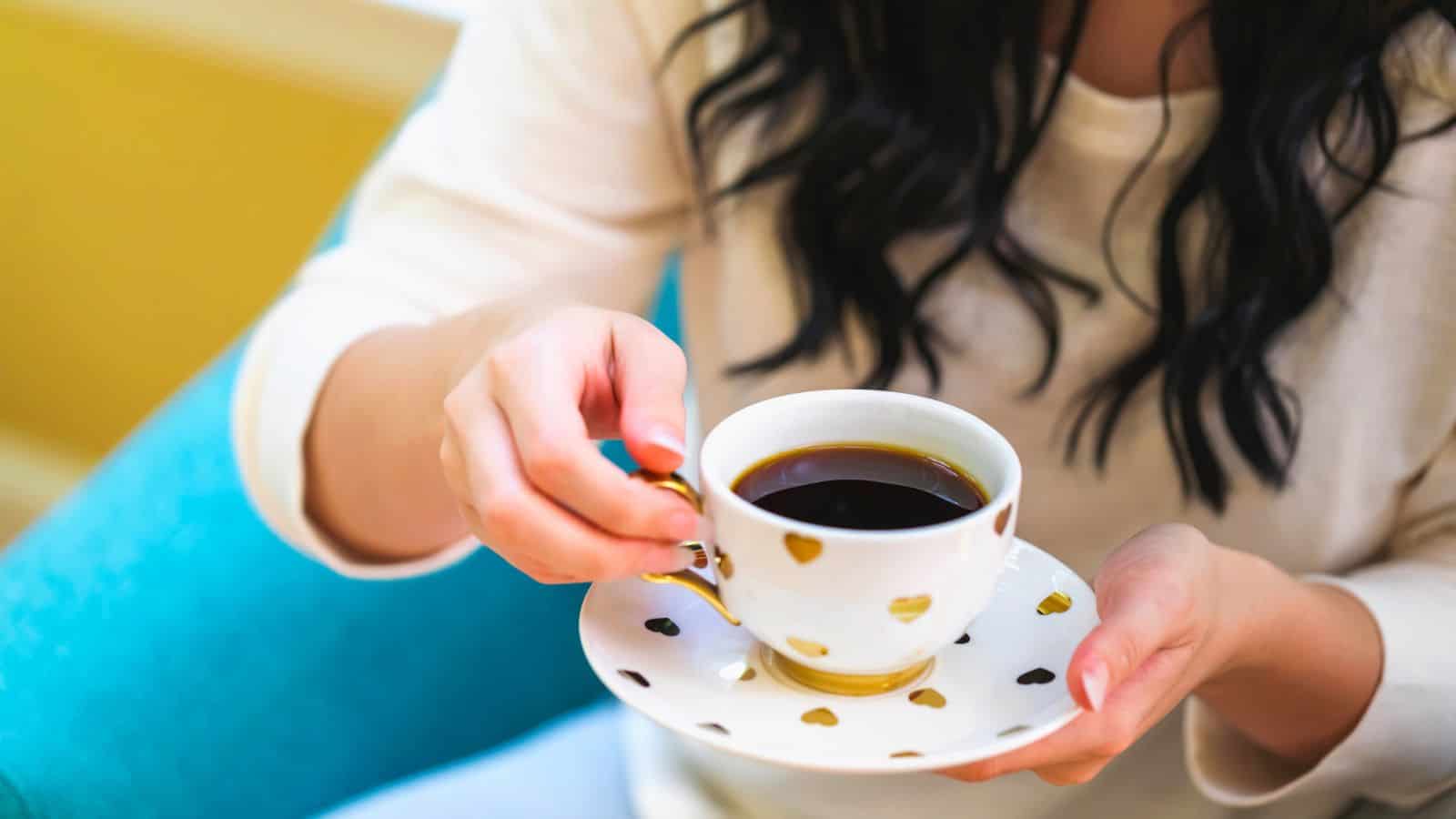 Woman holding a white cup with gold heart patterns, filled with coffee, sitting on a blue chair.