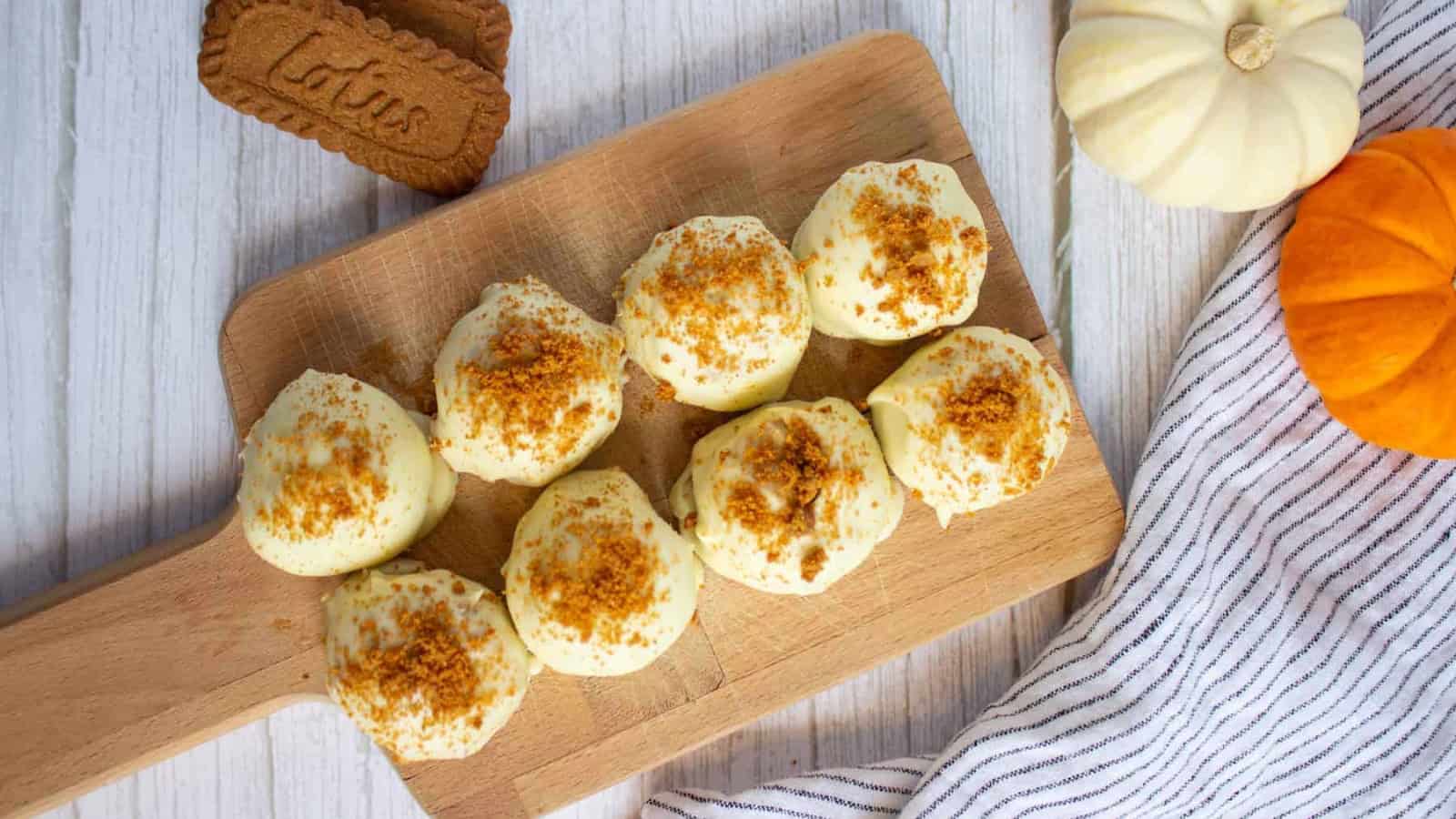 White chocolate pumpkin truffle topped with cookie crumbs, next to whole cookies on a white wooden surface.