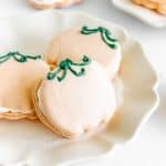Pumpkin Macarons shaped like pumpkins with green icing stems are displayed on a white plate.