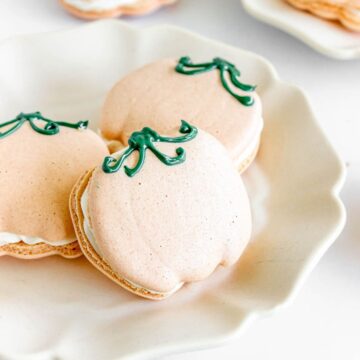 Pumpkin Macarons shaped like pumpkins with green icing stems are displayed on a white plate.