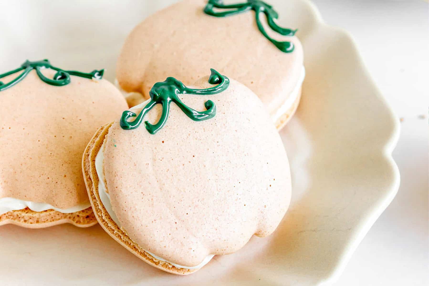 Pumpkin Macarons shaped like pumpkins with green icing stems sit beautifully on a white plate.