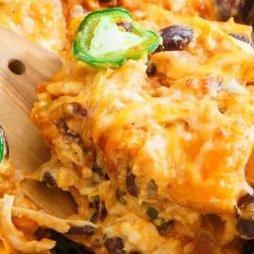 Cheesy casserole with black beans, shredded chicken, and sliced jalapeños being served with a wooden spatula.