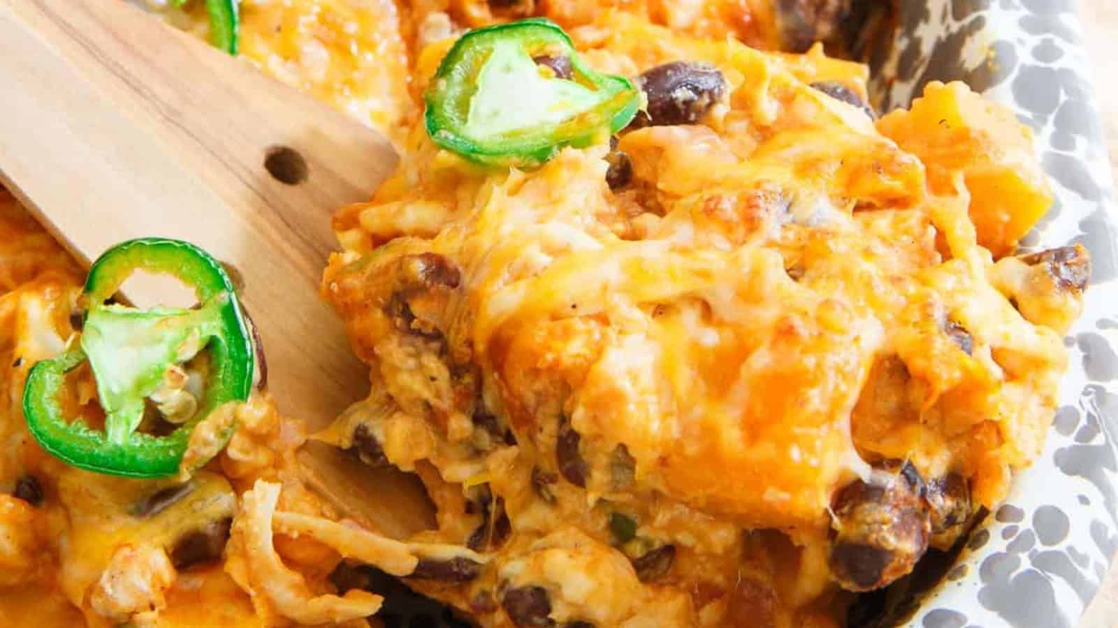 Cheesy casserole with black beans, shredded chicken, and sliced jalape&ntilde;os being served with a wooden spatula.