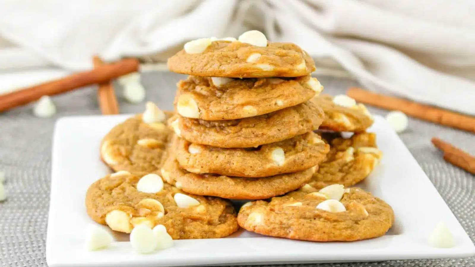 A stack of cookies with white chocolate chips on a white plate, surrounded by more chips and cinnamon sticks.