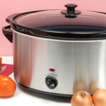 A stainless steel slow cooker with a black lid, next to carrots, onions, and a tomato on a pink background.
