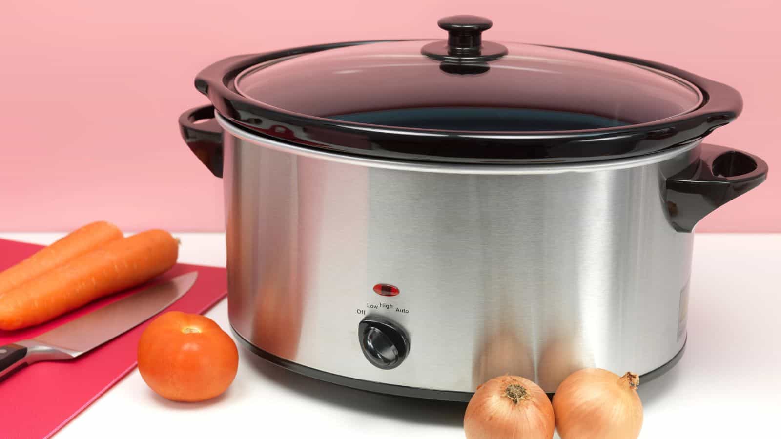 A stainless steel slow cooker with a black lid, next to carrots, onions, and a tomato on a pink background.