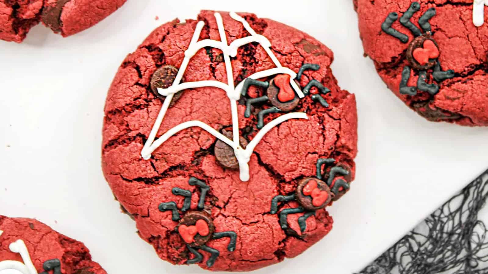 A red cookie decorated with white icing in a spiderweb pattern and black icing spiders with red details. Other similarly decorated cookies are partially visible around it.