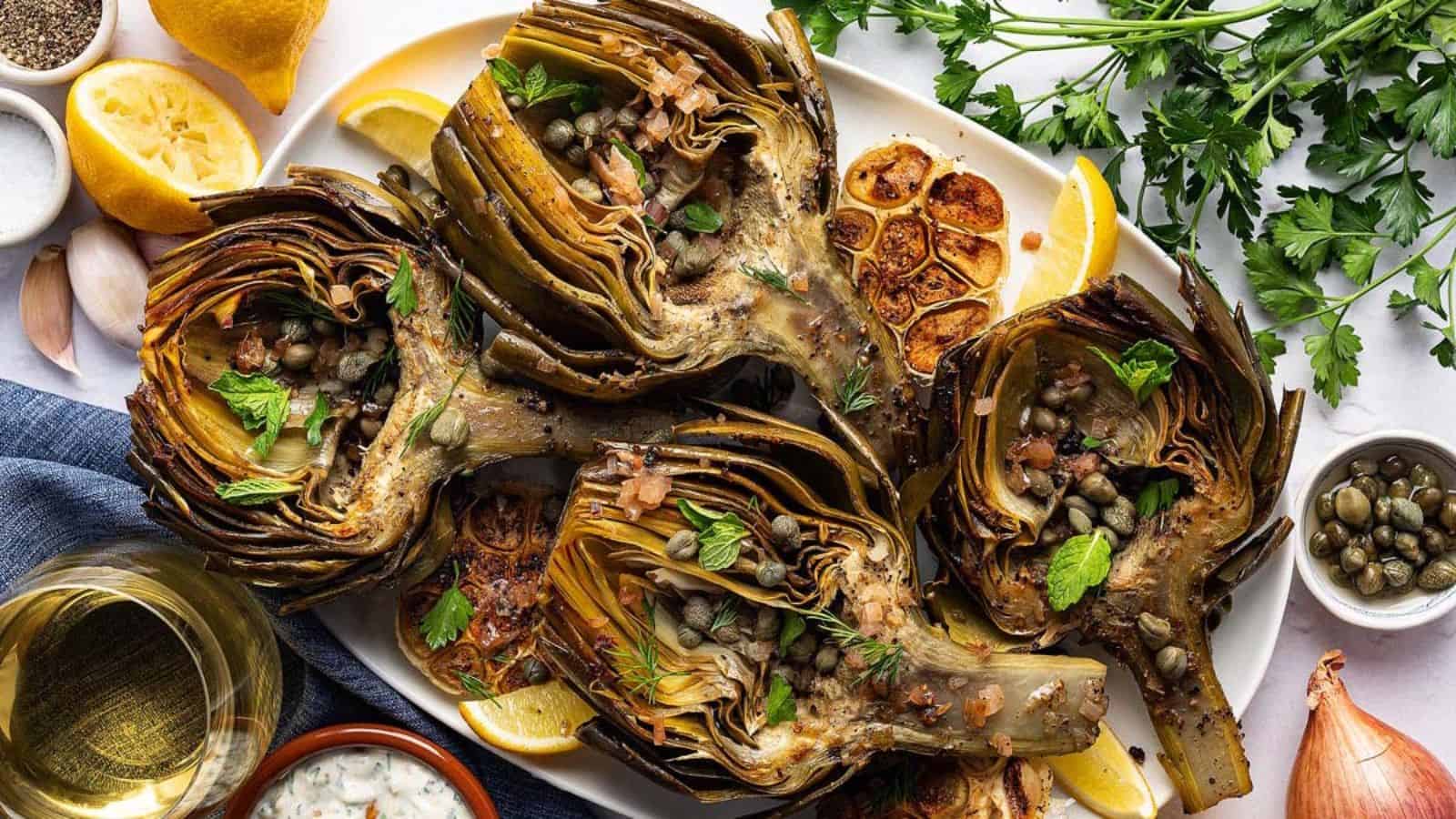 Grilled artichokes on a platter with lemon wedges, herbs, roasted garlic, and capers, with wine on the side.