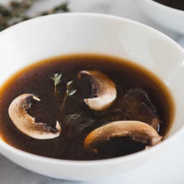A white bowl of dark mushroom broth with three mushroom slices and a sprig of thyme on top.