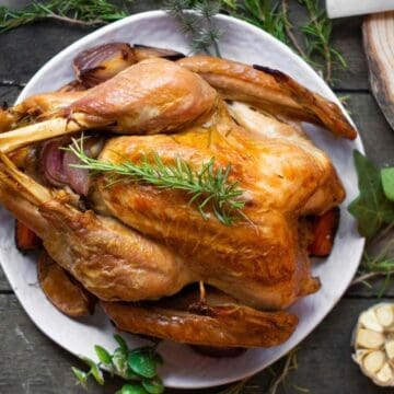 A roasted turkey on a white plate, garnished with rosemary, on a rustic wooden table.