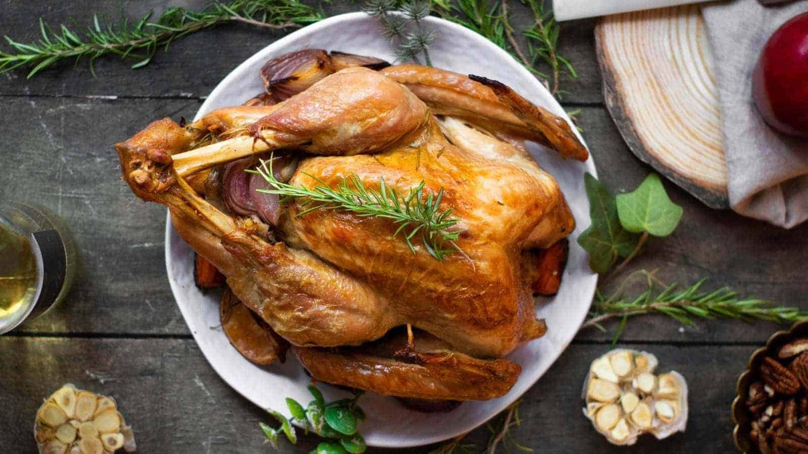 A roasted turkey on a white plate, garnished with rosemary, on a rustic wooden table.