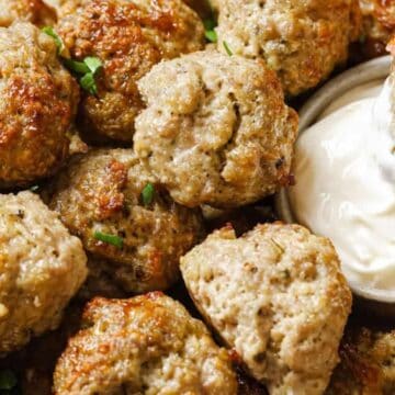 A hand dips a baked meatball into a bowl of creamy white sauce, surrounded by more meatballs on a plate.