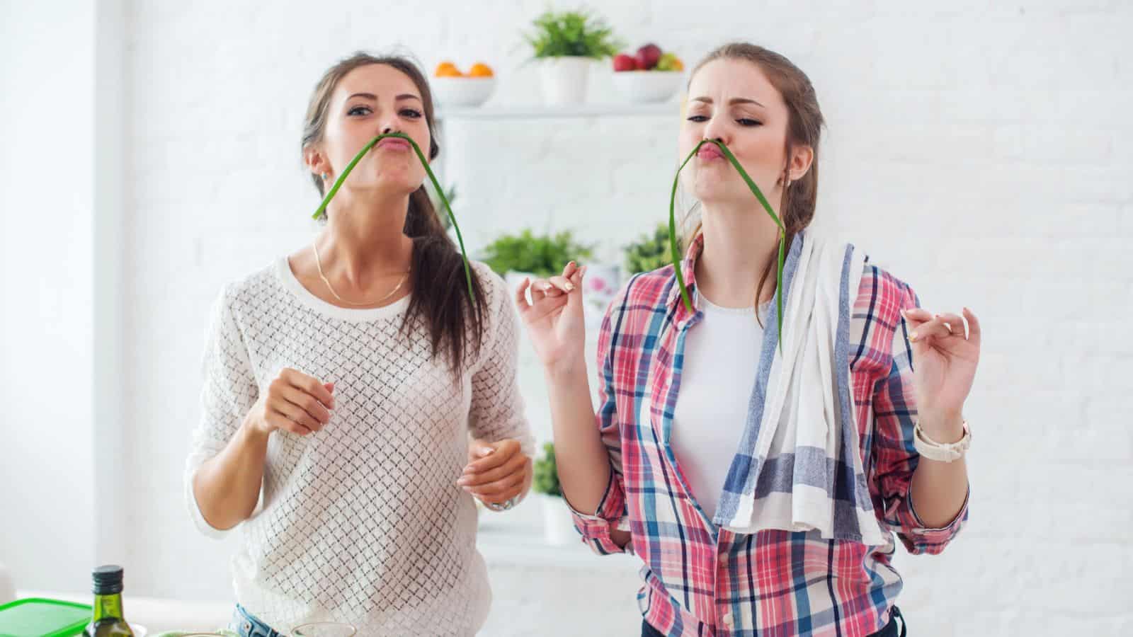 Two young women playfully hold green beans as mustaches while standing in a bright kitchen filled with essential cooking tools for beginners.