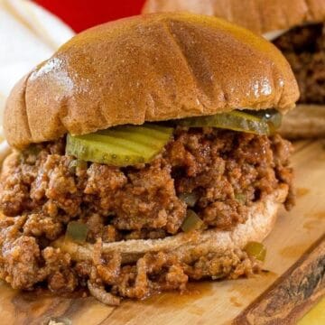 Two sloppy joe sandwiches with pickles on a wooden board, with a yellow checkered cloth in the background.