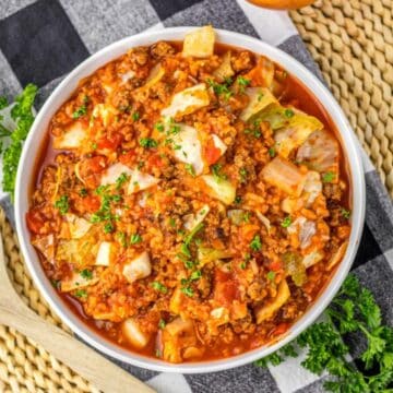 A bowl of hearty cabbage roll soup with ground meat and tomatoes, garnished with parsley, on a woven mat.