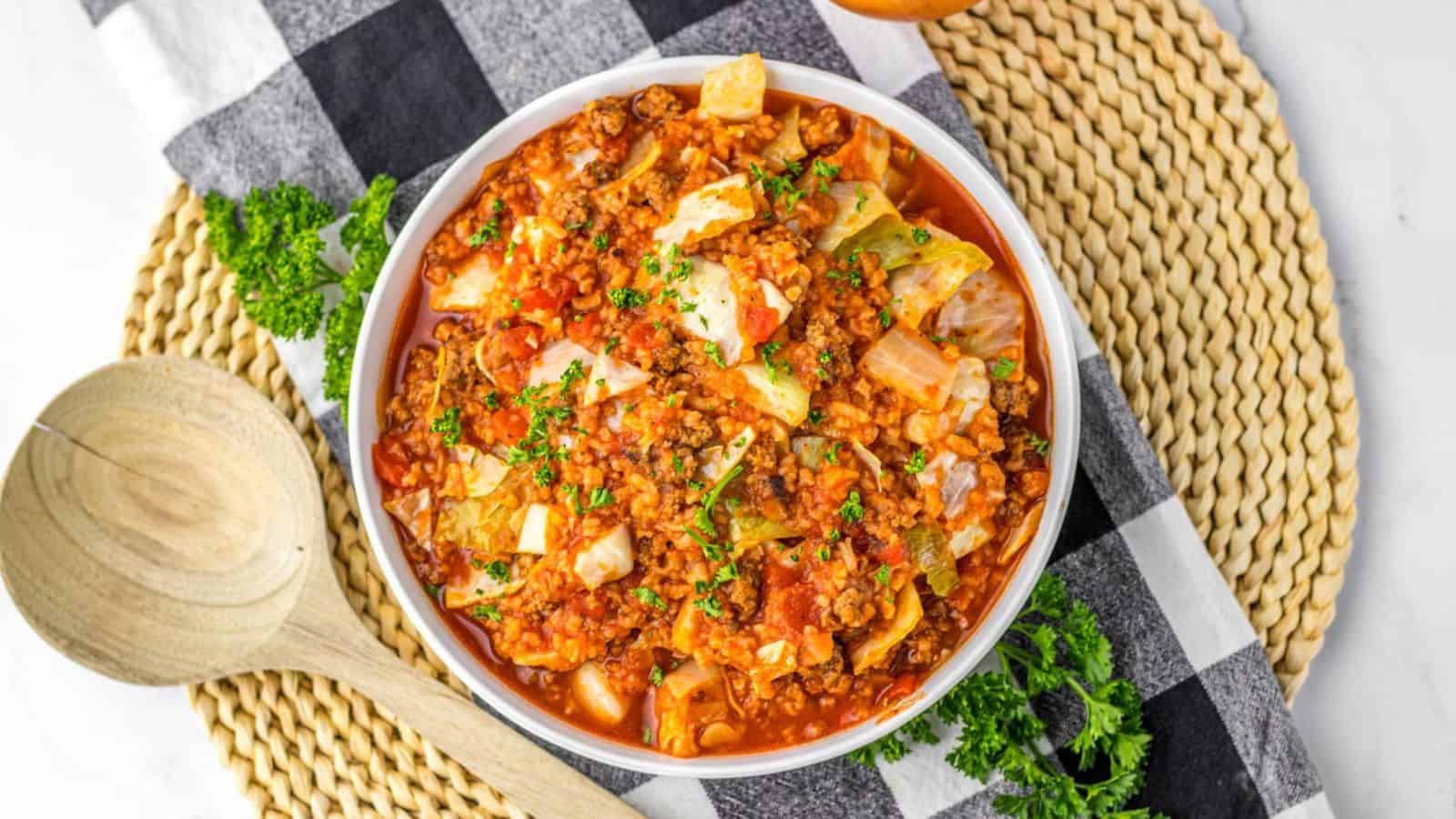 A bowl of hearty cabbage roll soup sits on a black and white checkered cloth atop a woven mat. The slow cooker soup is rich and chunky, filled with tender cabbage, ground meat, and tomatoes, and garnished with fresh parsley. Wooden utensils are placed beside the bowl.