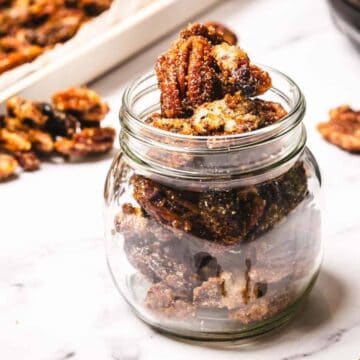 A glass jar filled with candied pecans, with more pecans on a tray and scattered on a white surface nearby.