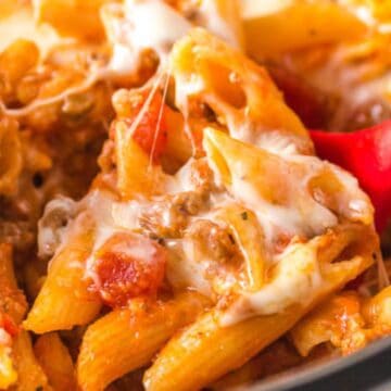 Cheesy baked penne pasta with ground meat and tomato sauce, being stirred with a red spoon.