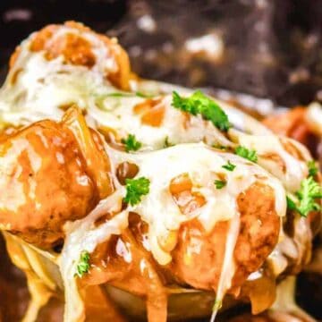 A ladle lifts cheesy meatballs topped with melted cheese and parsley in a rich brown sauce.