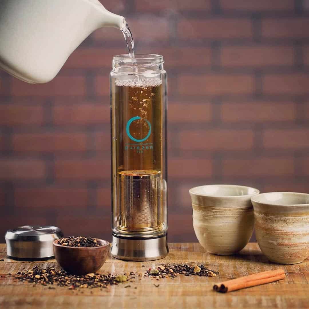 Glass tea infuser bottle being filled with hot water, surrounded by two cups and loose tea leaves on a table.