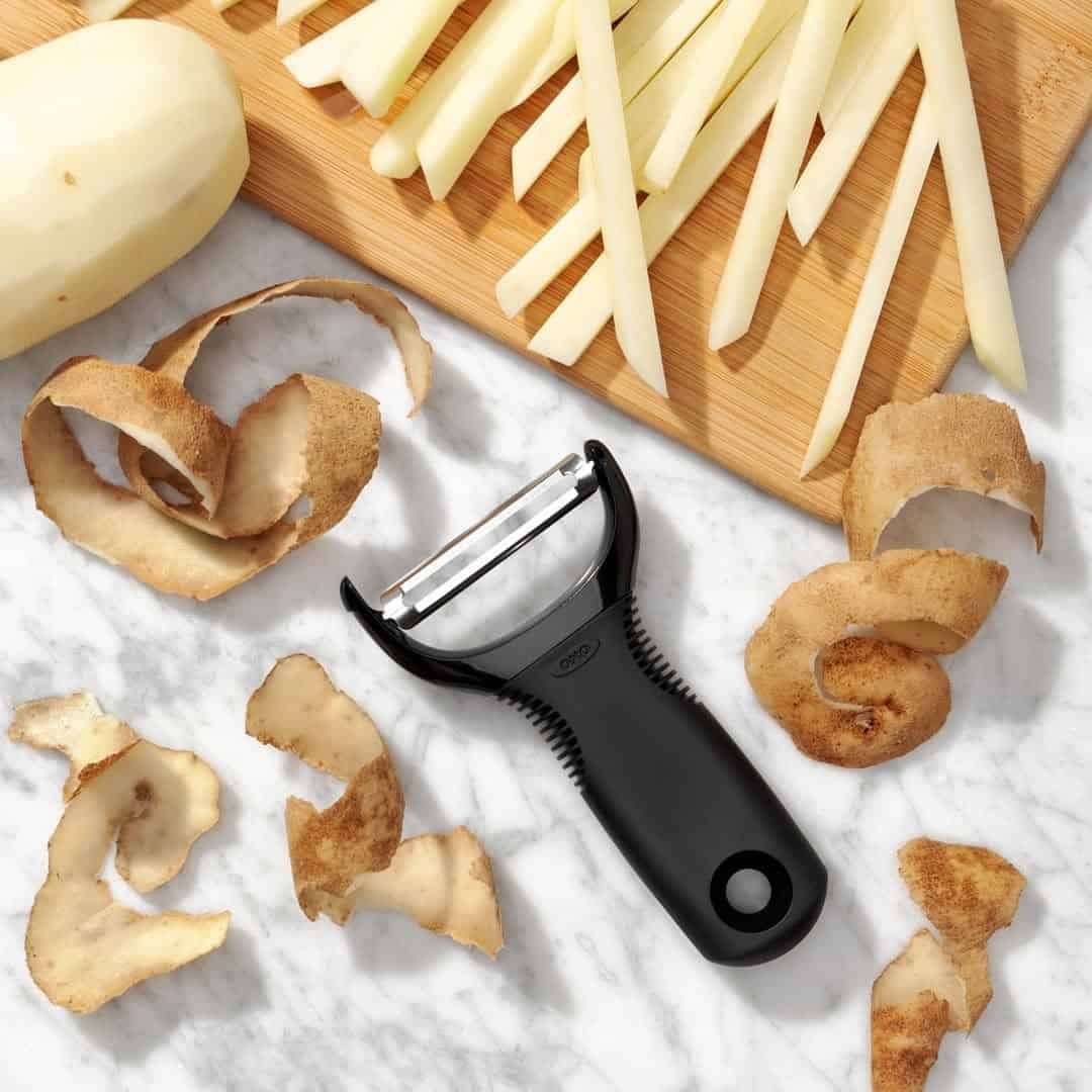 A black peeler, potato peelings, a peeled potato, and sliced potatoes on a cutting board-essential cooking tools for beginners making meal prep easy and efficient.