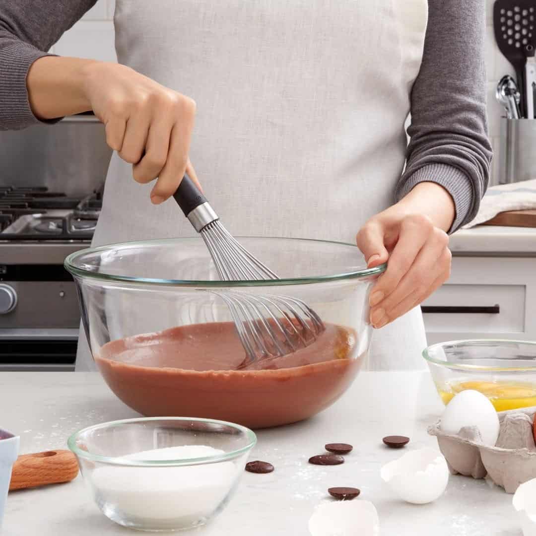 Person whisking chocolate batter in a glass bowl with essential cooking tools for beginners, surrounded by baking ingredients on a kitchen counter.