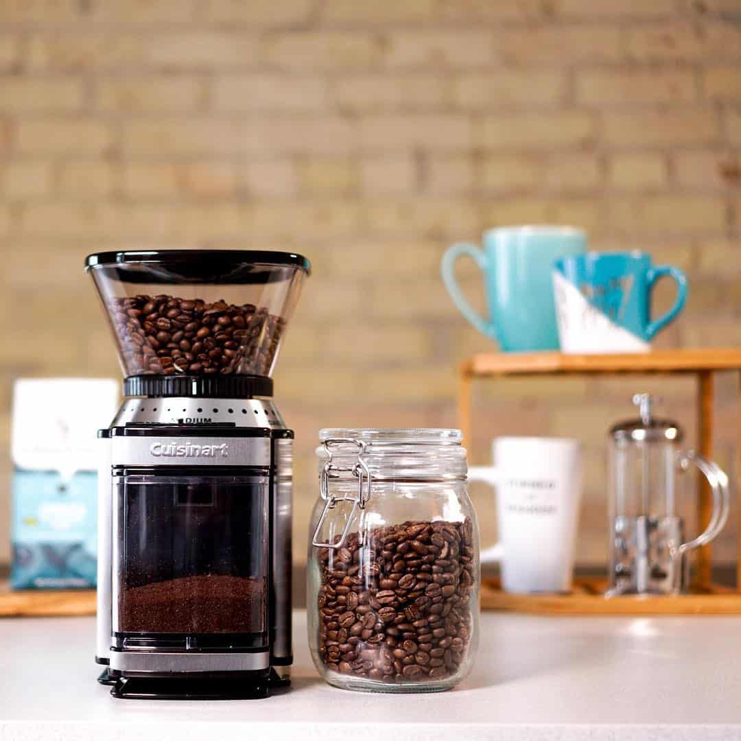 Coffee grinder with beans, a jar of coffee beans, mugs, and a French press on a counter.