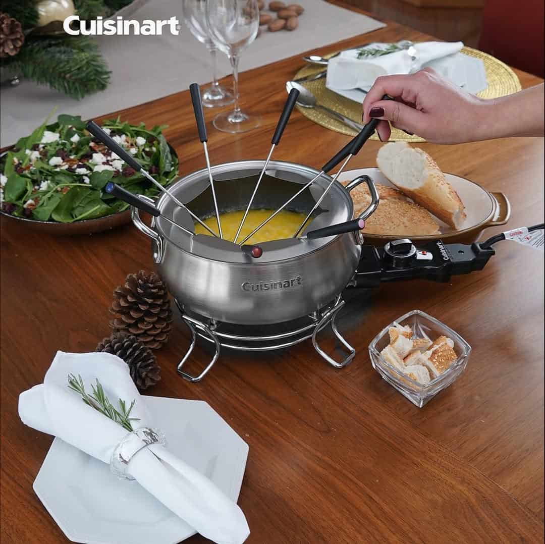 A hand dips a fondue fork into a Cuisinart fondue pot on a set dining table with bread and salad—perfect for gifts for cheese lovers.