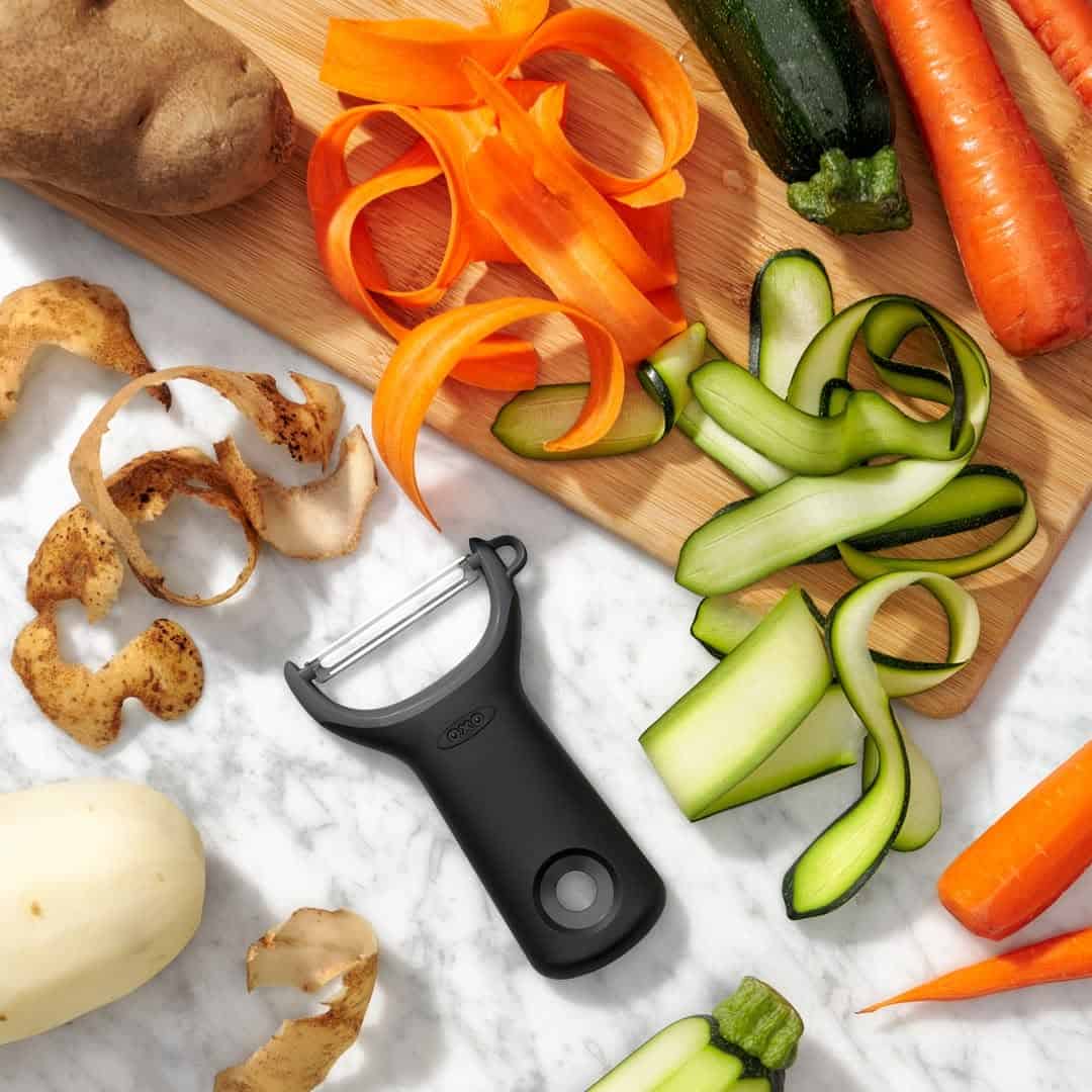 A black vegetable peeler with peelings and sliced carrots, zucchinis, and potatoes on a marble counter—an essential tool and a thoughtful idea for gifts for cheese lovers who enjoy fresh, home-cooked meals.