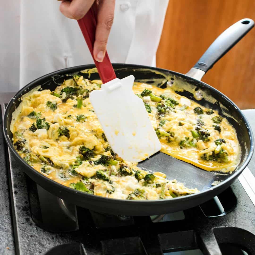 A person cooking scrambled eggs with broccoli in a skillet, using a spatula—one of the essential cooking tools for beginners—to stir on a stovetop.