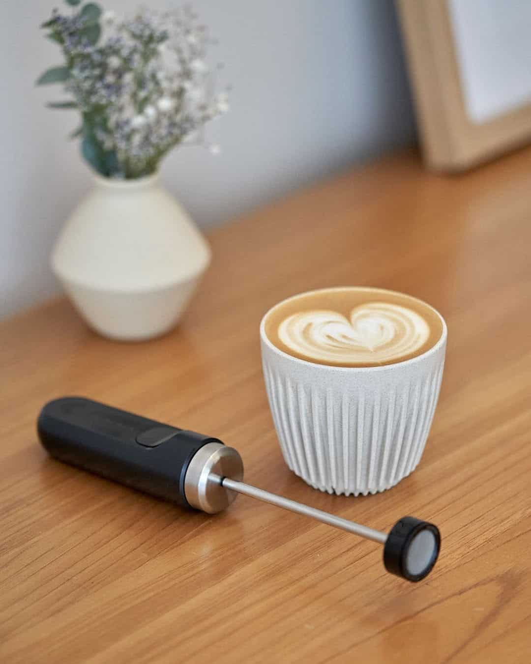 A cup of latte with heart art, a milk frother, and a vase of flowers on a wooden table.