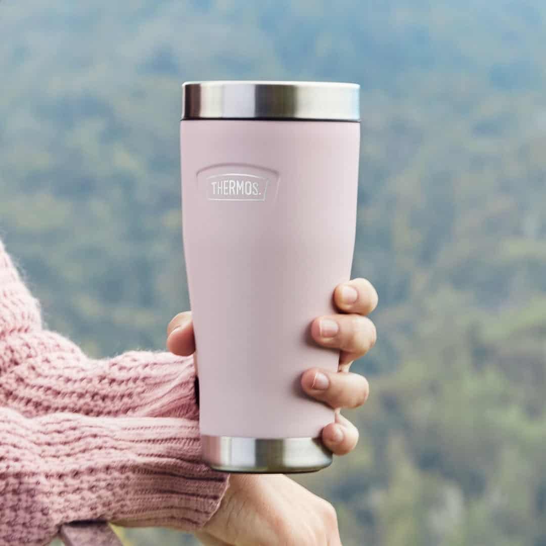 A hand holds a pink Thermos travel mug outdoors with a blurred green background.