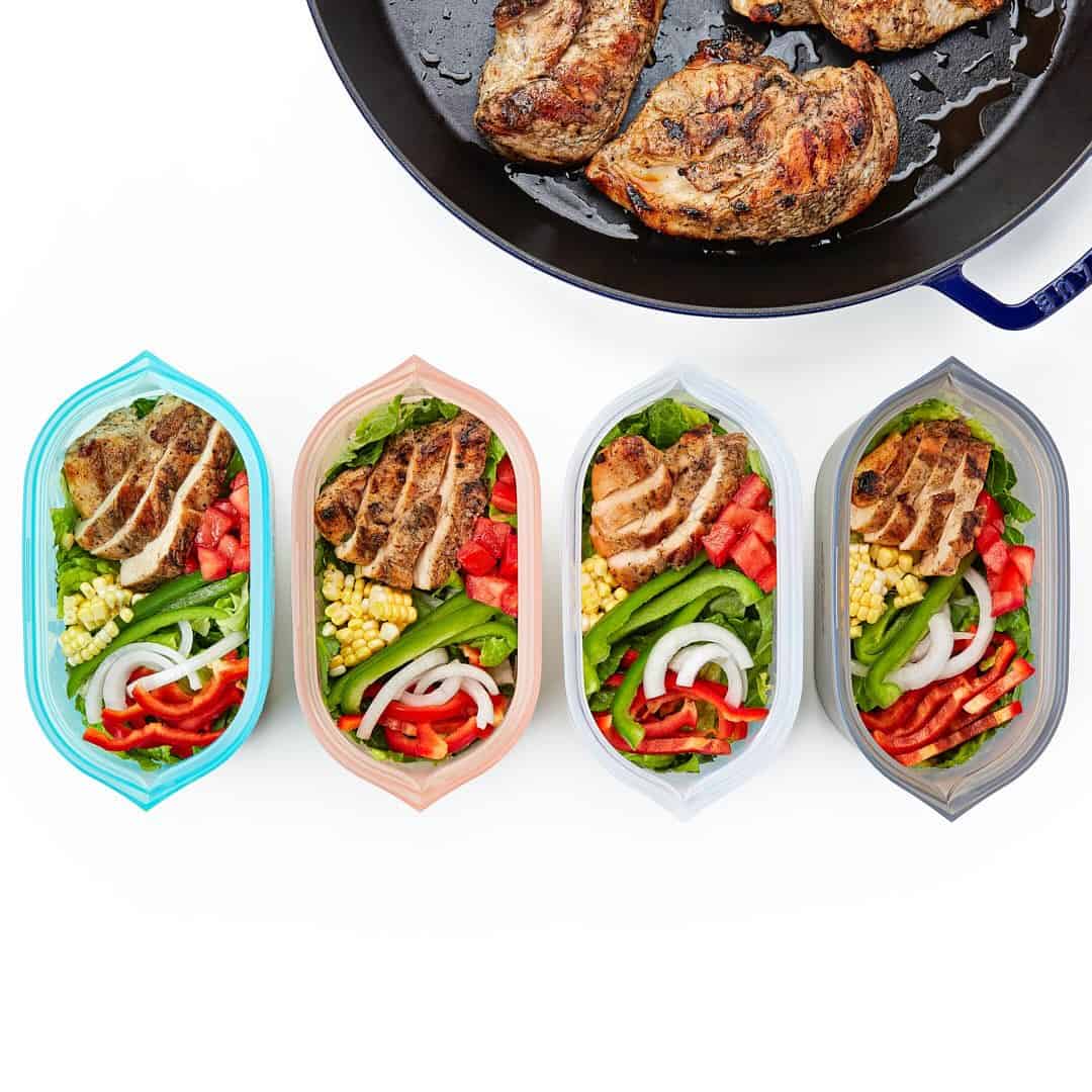 Four containers of salad with grilled chicken, corn, peppers, onions, and greens sit beside a pan of grilled meat-perfect food storage gifts for meal prepping or sharing healthy lunches.