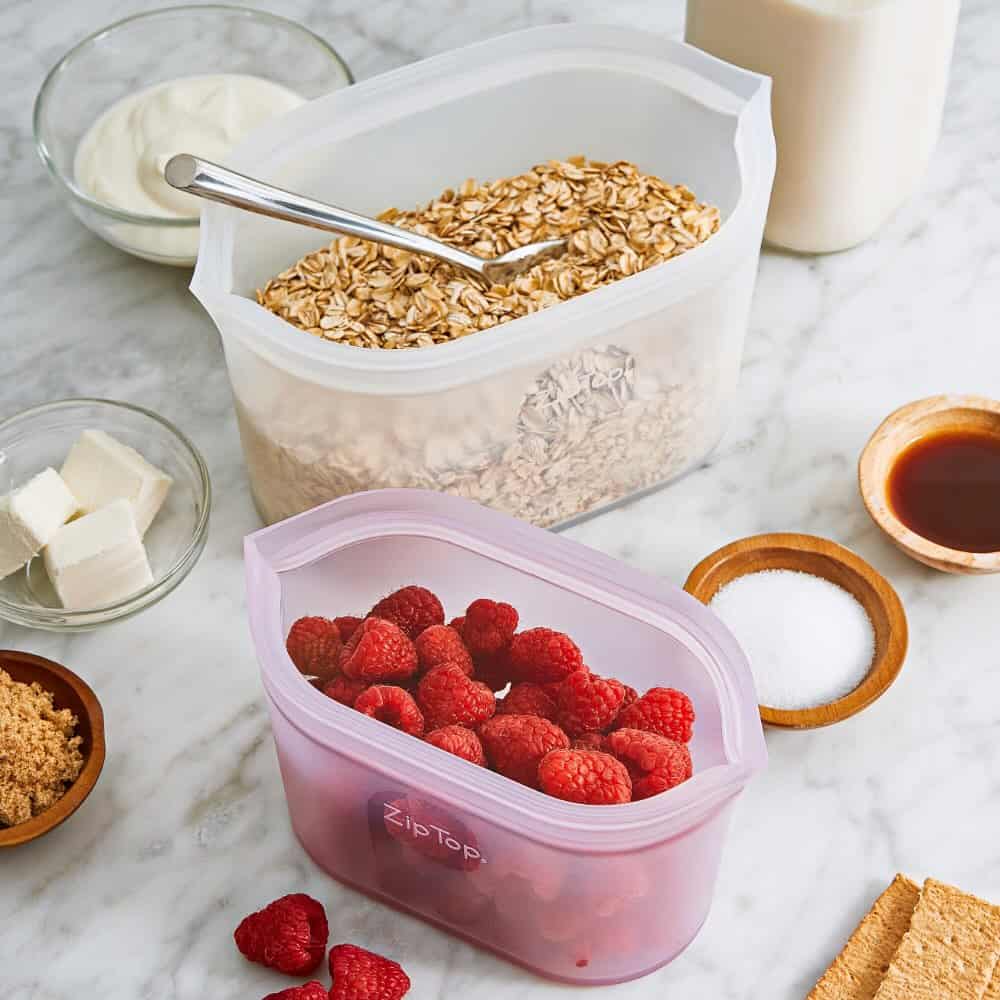 Two reusable silicone bags filled with oats and raspberries sit on a marble counter with baking ingredients-practical food storage gifts for any kitchen.